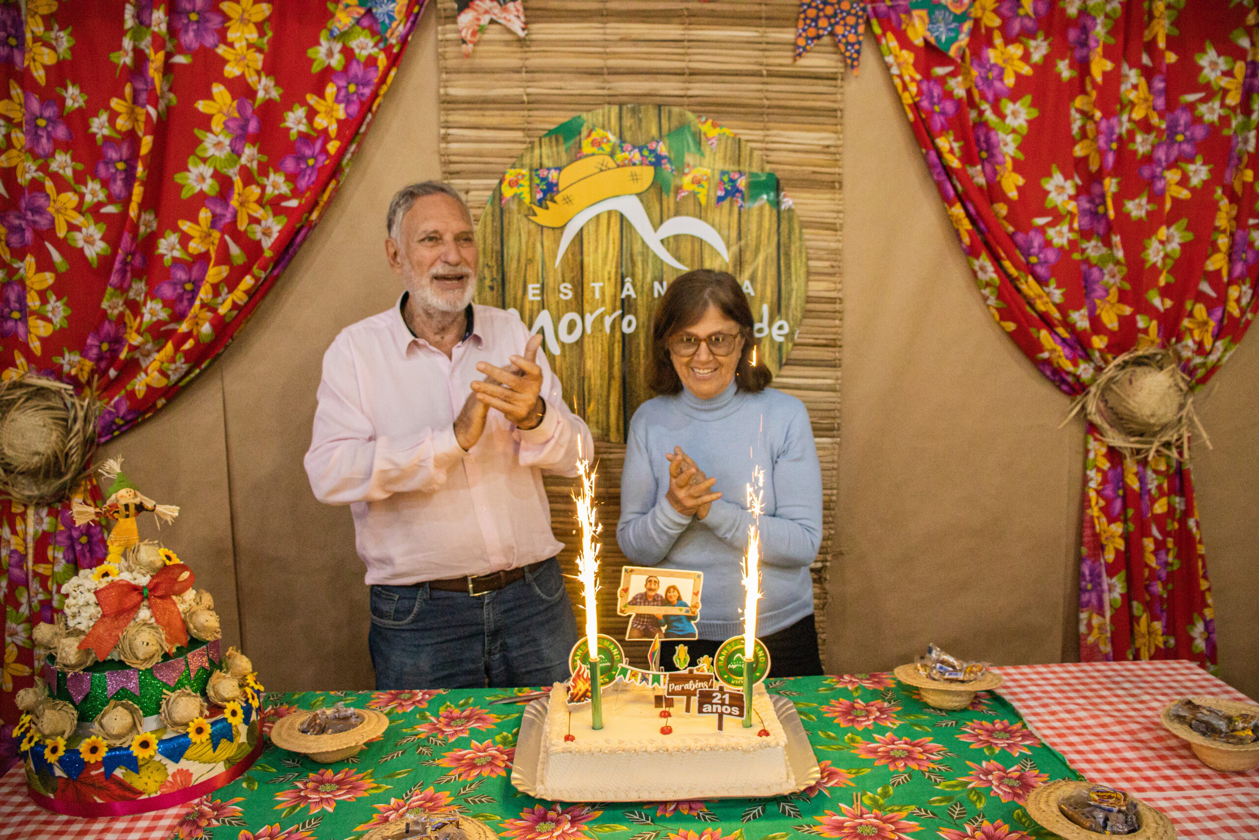 Dr. Juan M. Urbinati e Beatriz Urbinati em parabéns aos 21 anos do Estância Morro Grande. Fundo de cortinas vermelhas com florais amarelos, esteira com o logo HEMG adornado com chapéu junino e bandeirolas. Mesa com bolo fake de pipoca e um bolo real com toppers personalizados com inscrições de 